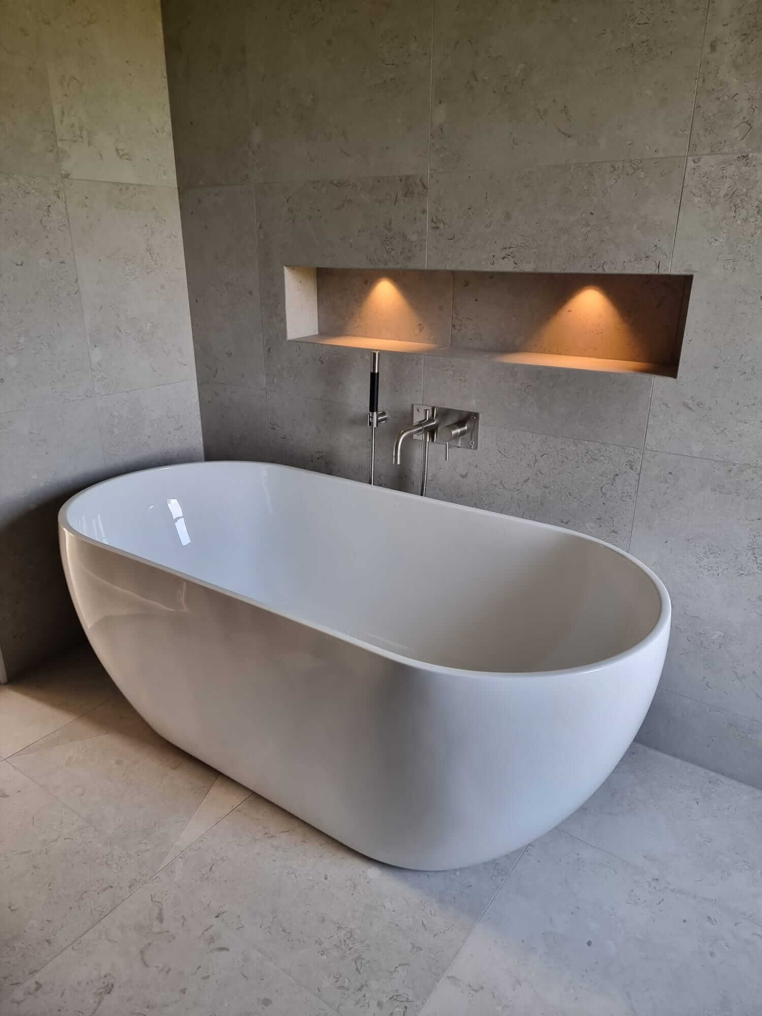 Freestanding white bathtub in elegant beige bathroom with wall lighting.
