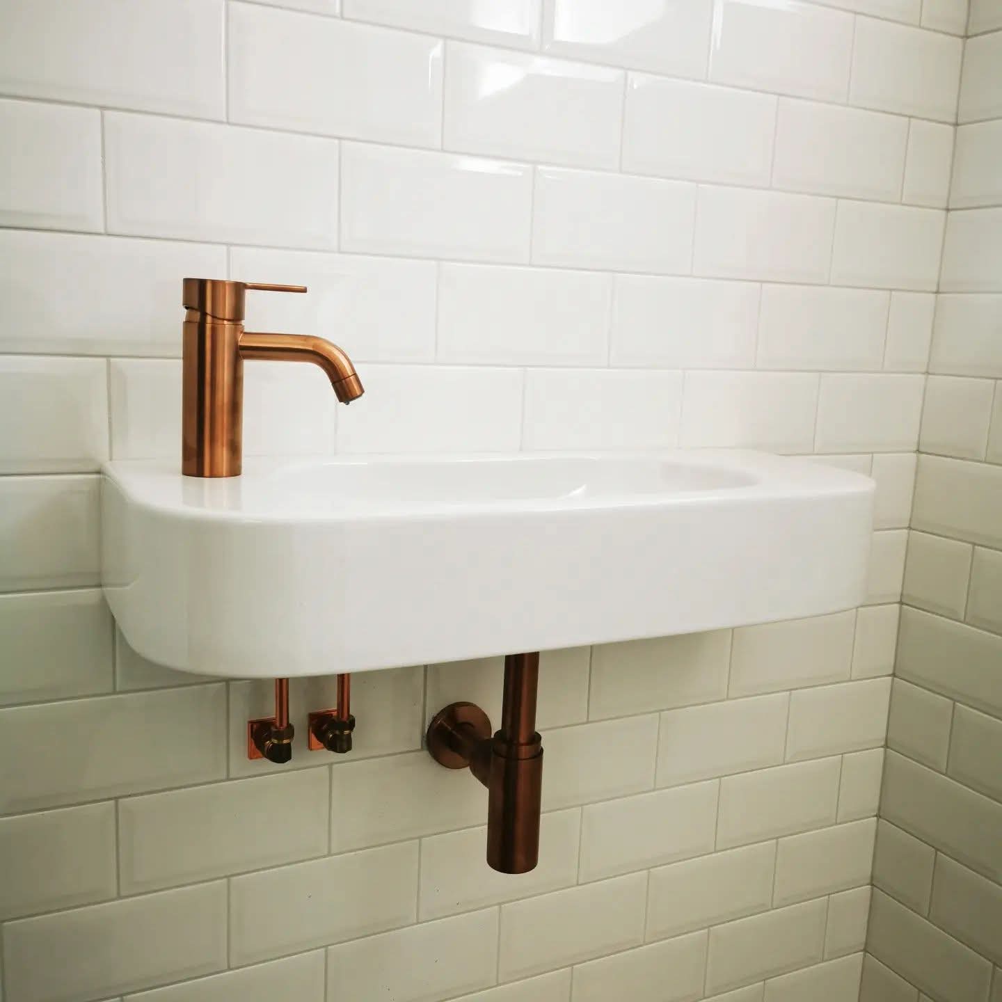 Small white sink with copper faucet and visible copper pipes on white tiled wall.